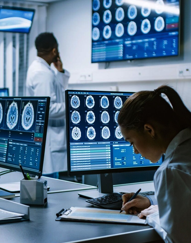 Lekarka analizuje wyniki tomografii komputerowej mózgu na monitorze, notując dane na arkuszu, w tle drugi lekarz rozmawia przez telefon w sterylnym pomieszczeniu.