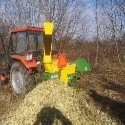 Czerwony traktor z otwartymi drzwiami kabiny i ż&oacute;łto-zielony rębak do gałęzi, pracujący na polu z widoczną stertą zrębk&oacute;w drzewnych w tle drzew i krzew&oacute;w.