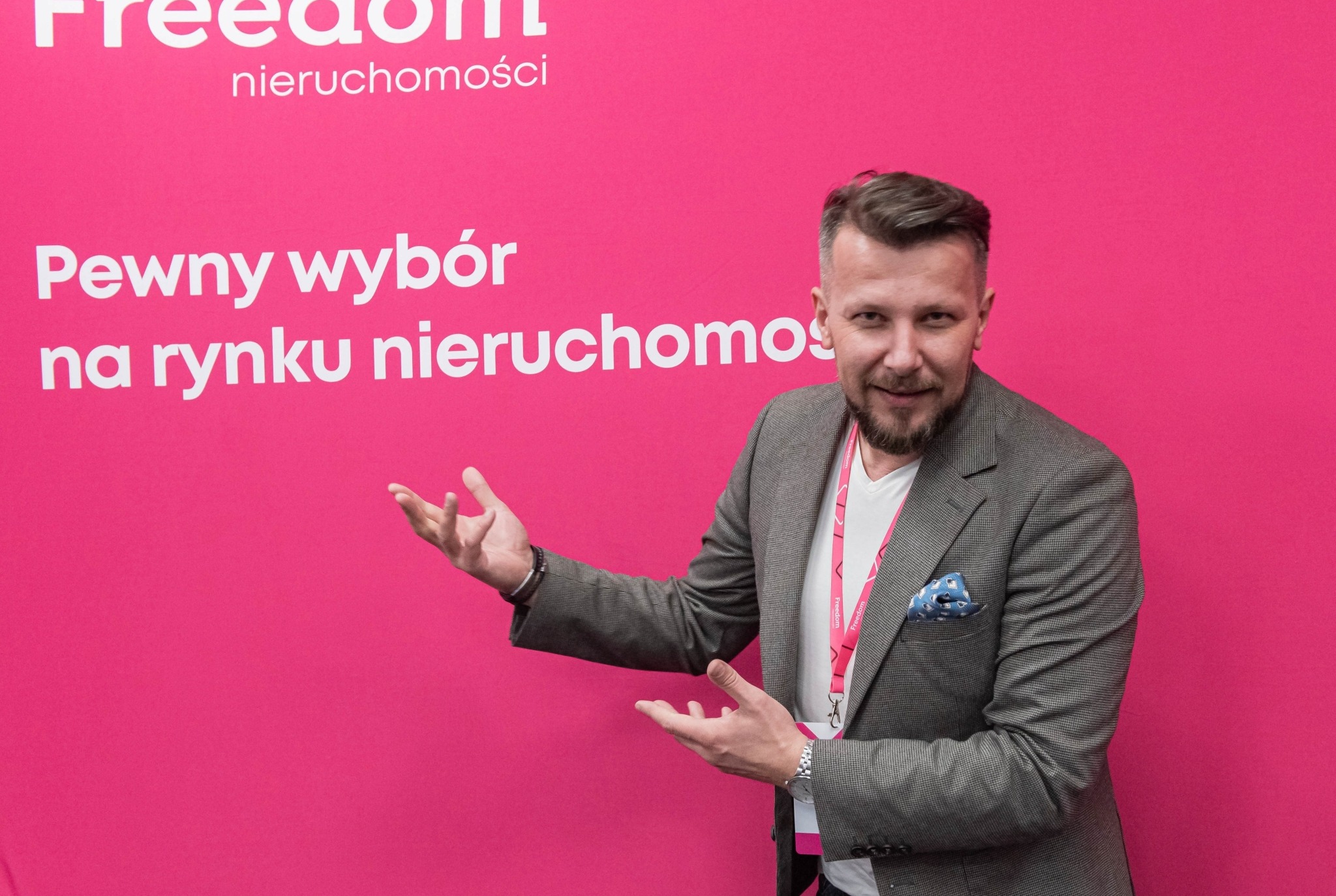 Mężczyzna w marynarce gestykuluje na tle różowej ściany z logo 'Freedom nieruchomości' i napisem 'Pewny wybór na rynku nieruchomości'.
