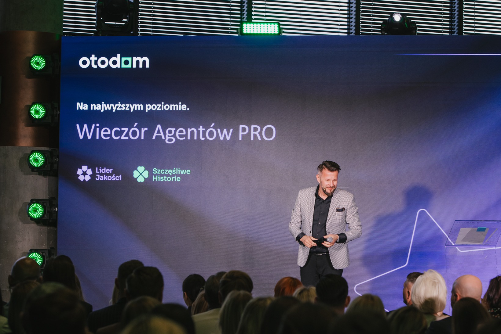 Prezentacja 'Wieczór Agentów PRO' na tle podświetlonej sceny z logo Otodom. Prelegent w marynarce i publiczność. Wydarzenie branżowe dla agentów nieruchomości.