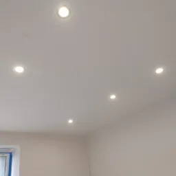 Biały sufit z zamontowanymi pięcioma okrągłymi lampami LED, widoczny fragment okna z niebieską folią ochronną, świeżo pomalowane ściany w jasnym odcieniu beżu.