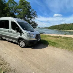 Srebrny bus Ford Transit zaparkowany na polnej drodze z widokiem na jezioro i las w tle, błękitne niebo z chmurami.