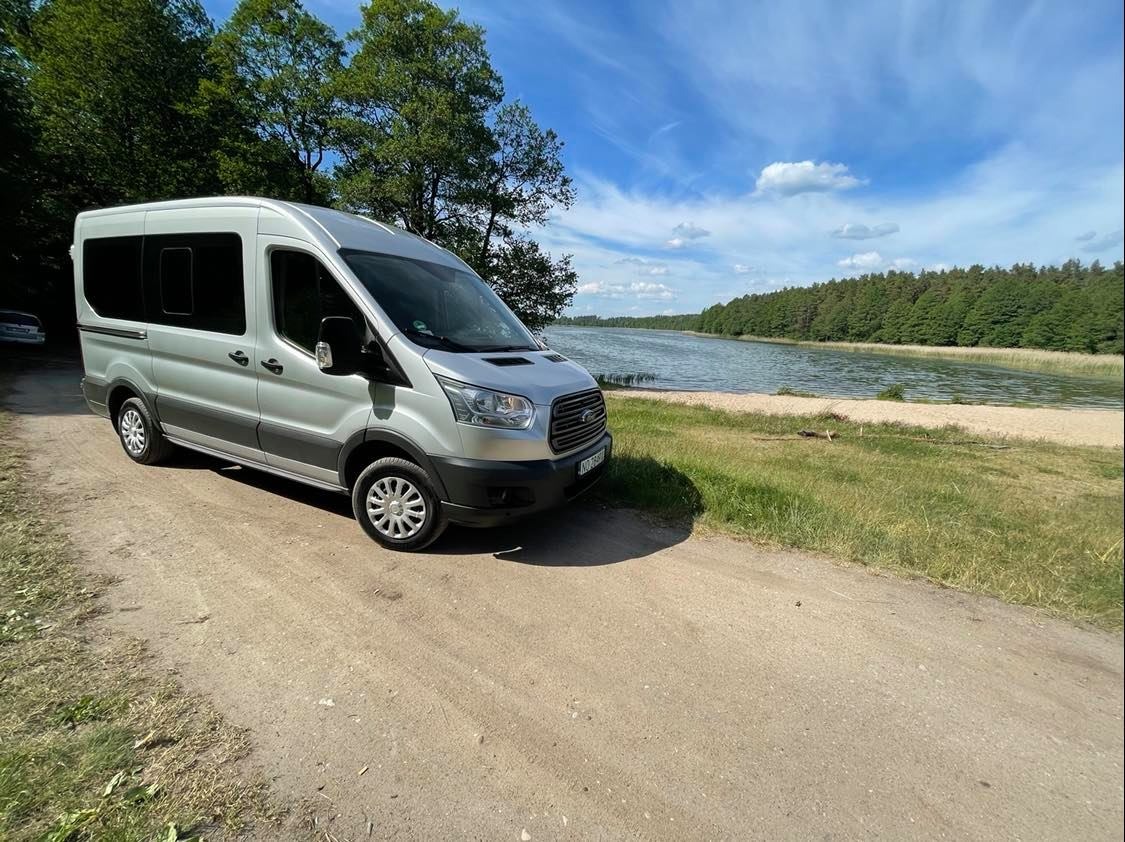 Srebrny bus Ford Transit zaparkowany na polnej drodze z widokiem na jezioro i las w tle, błękitne niebo z chmurami.
