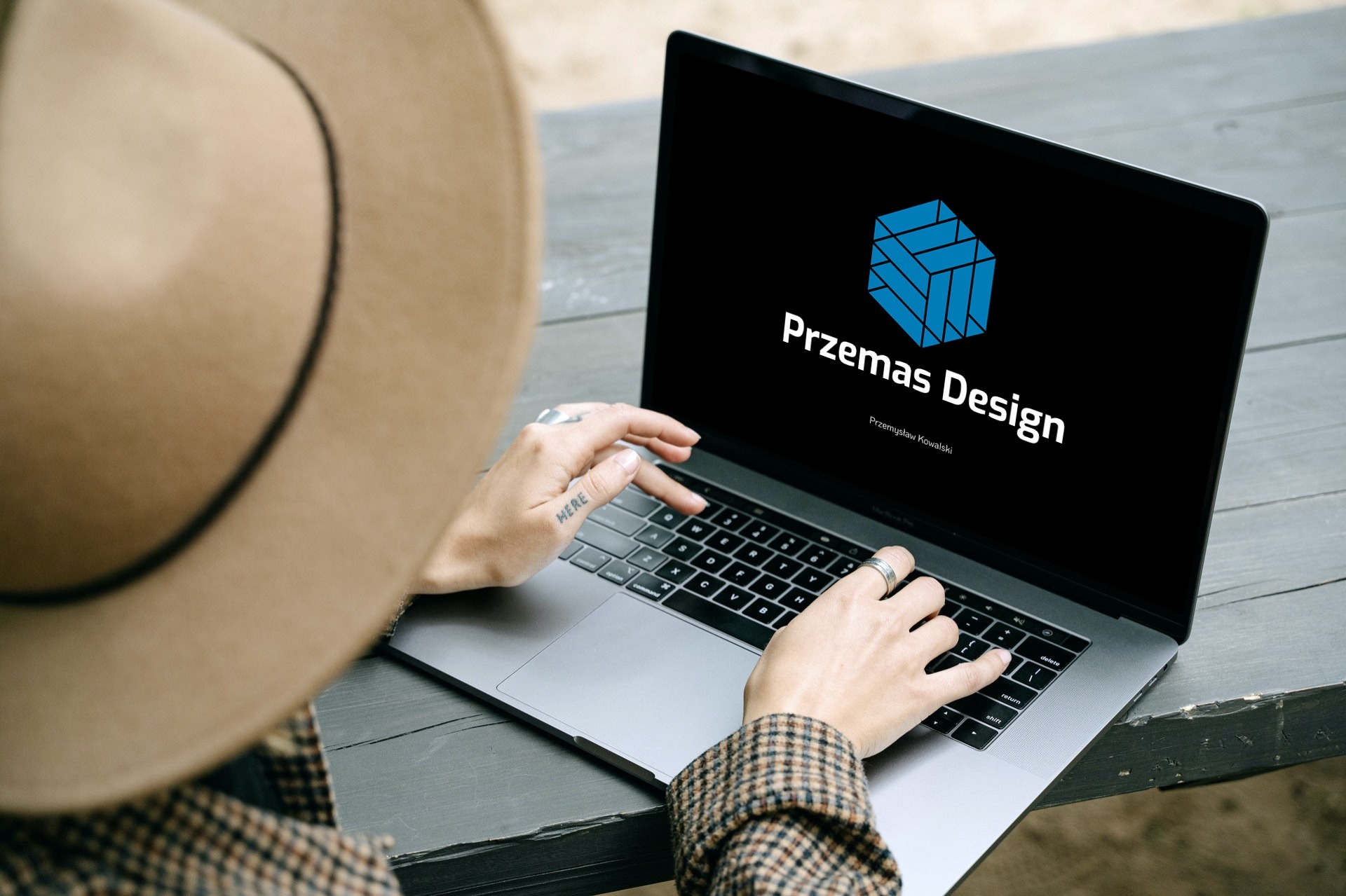 Osoba w kapeluszu i kraciastej marynarce pracuje na laptopie, na ekranie widoczne logo i nazwa 'Przemas Design' na czarnym tle.