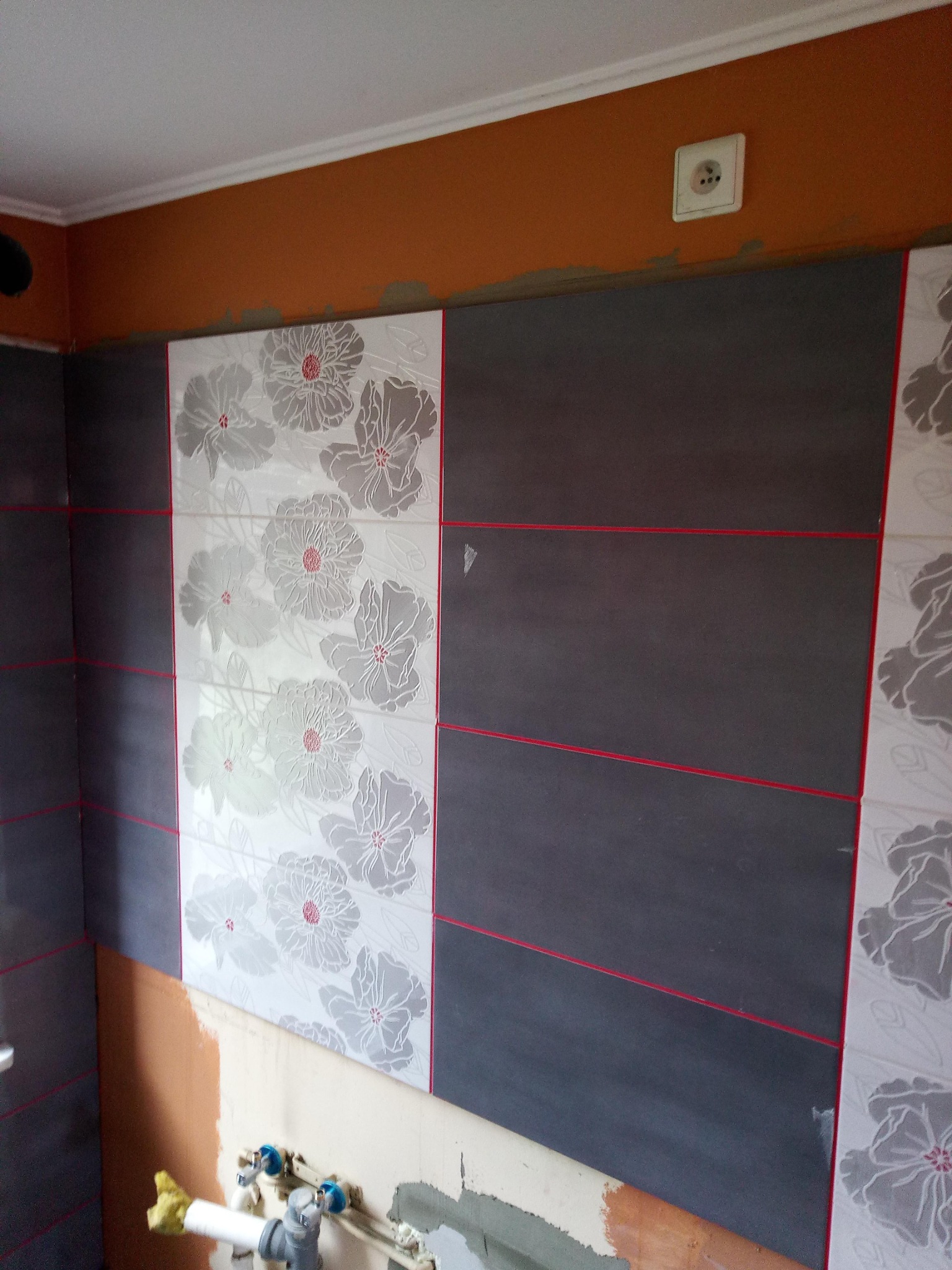 Fragment ściany w trakcie układania płytek w łazience, widoczne szare płytki z czerwonymi fugami oraz białe płytki z motywem kwiatowym, poniżej niezabezpieczone rury hydrauliczne i fragment ściany...