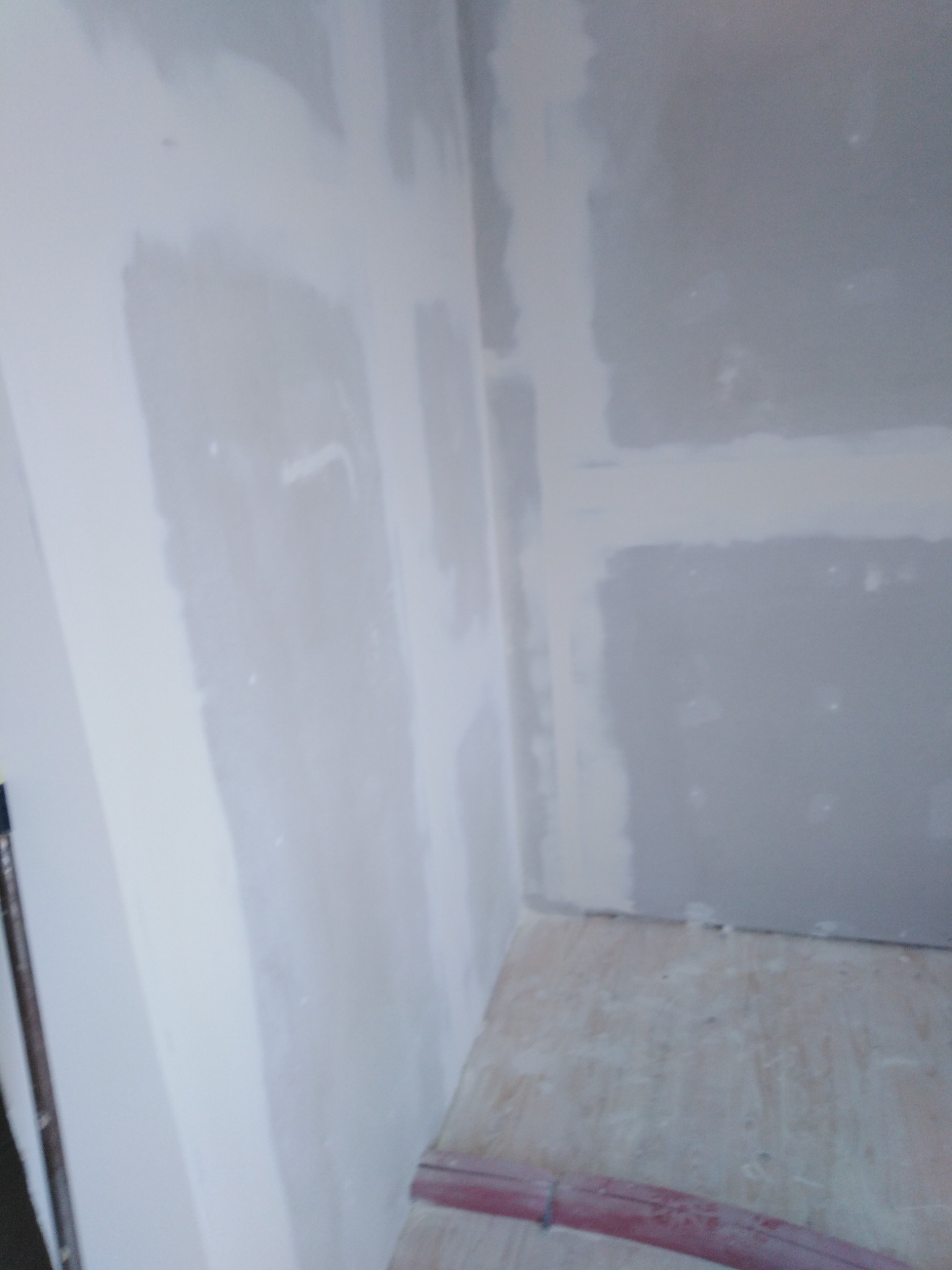 Niedokończone ściany z widocznymi łączeniami płyt i śladami szpachlowania, surowe drewniane podłoże z resztkami pyłu budowlanego, czerwony przewód na podłodze