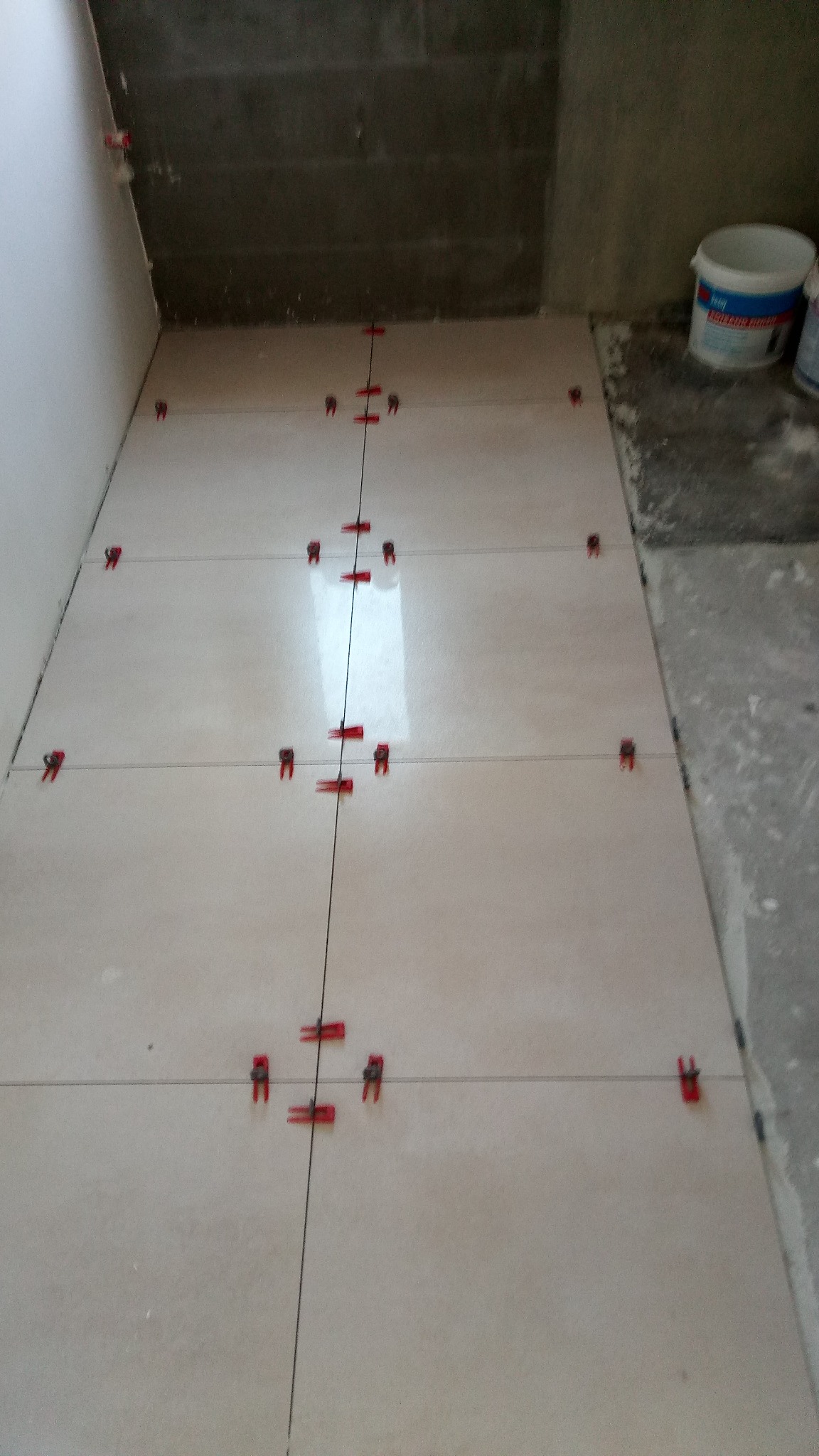 Układanie jasnych płytek podłogowych z użyciem czerwonych klipsów poziomujących i sznurka, widoczny fragment surowej ściany i wiaderka z zaprawą w tle.