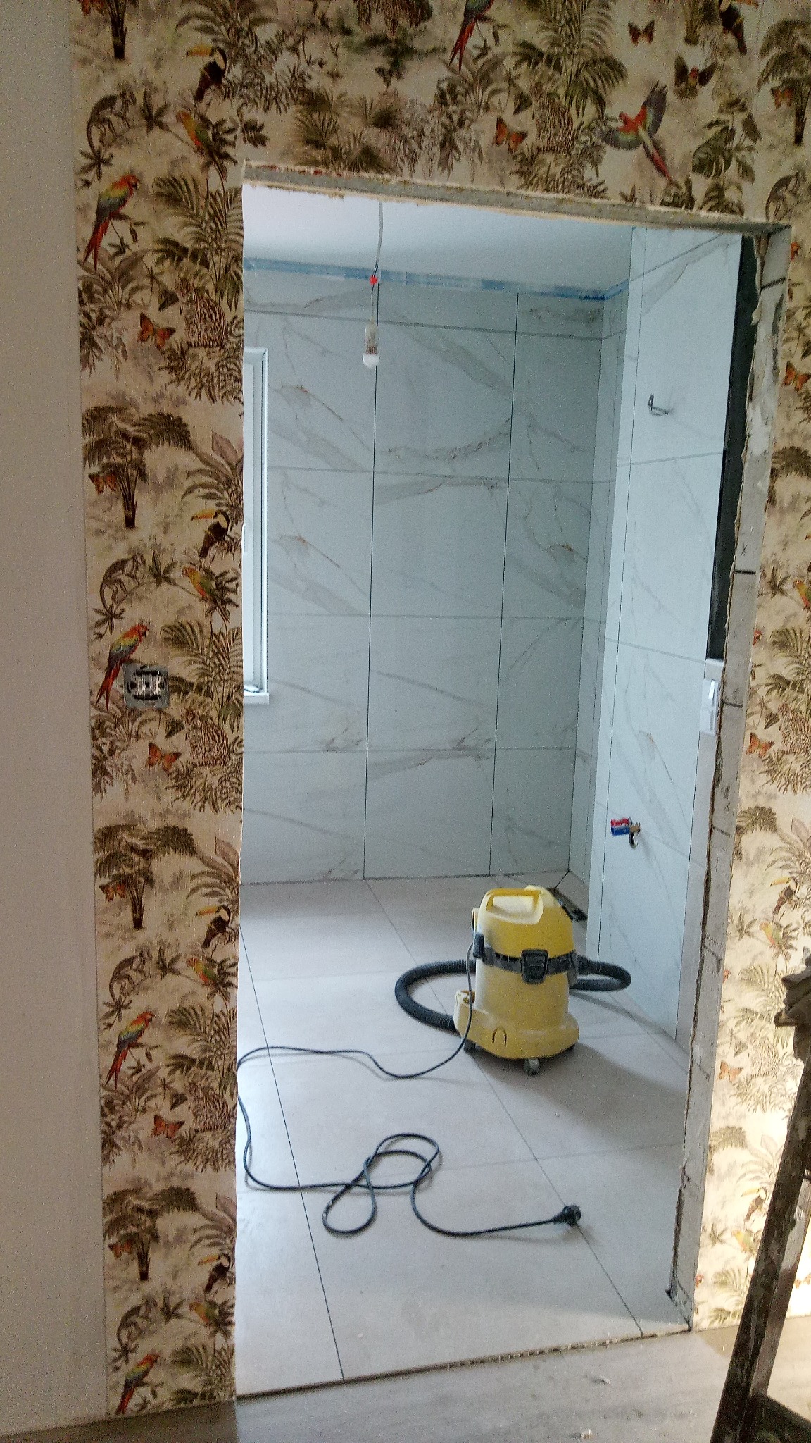 Łazienka w trakcie wykańczania: białe płytki imitujące marmur na ścianach, tapeta w tropikalne wzory na ścianie zewnętrznej, żółty odkurzacz przemysłowy na podłodze.
