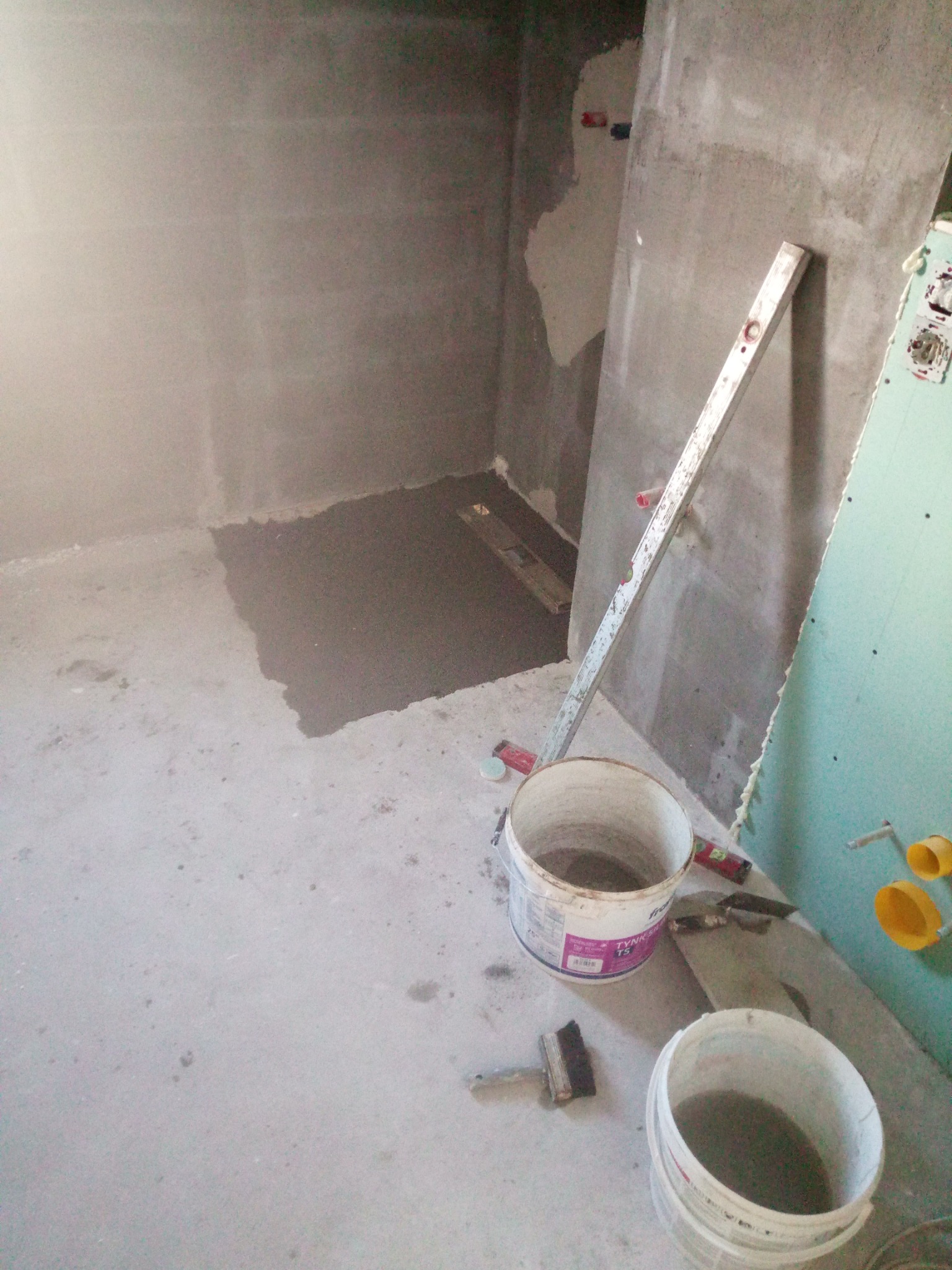 Surowy stan łazienki w trakcie remontu: szare ściany, częściowo wylany beton na podłodze, poziomica oparta o ścianę, dwa wiadra z zaprawą, pędzel i płyta gipsowa z instalacją elektryczną.