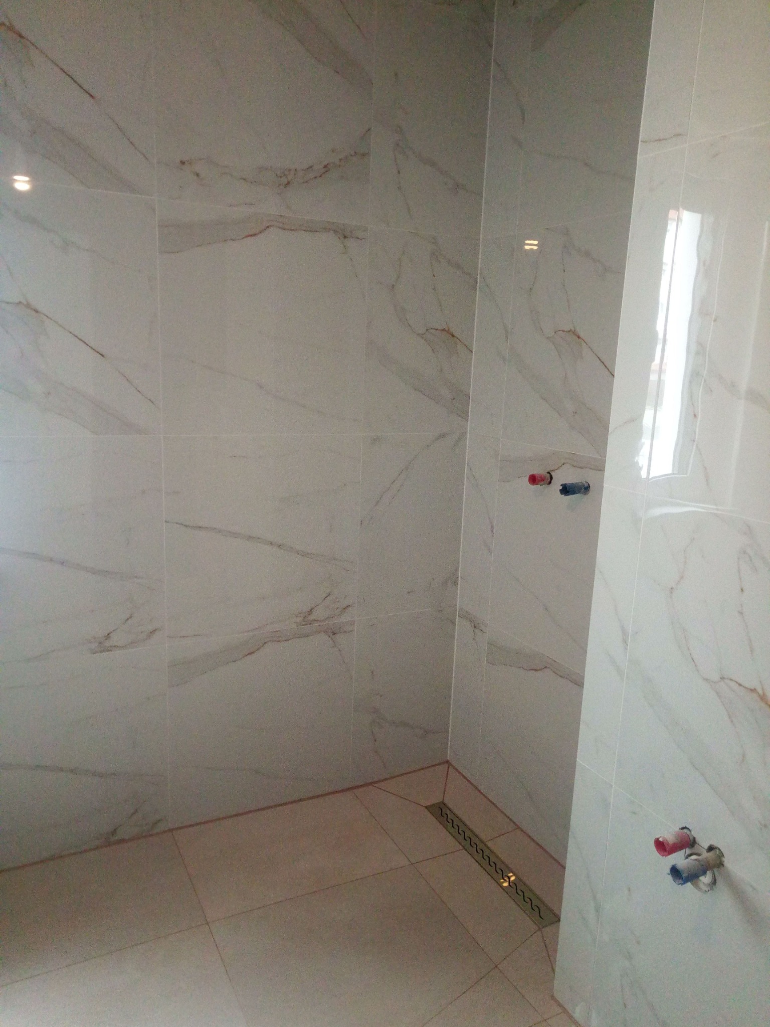 Wykończona łazienka z białymi płytkami imitującymi marmur, widoczny odpływ liniowy w podłodze i wystające przyłącza wodne ze ścian.