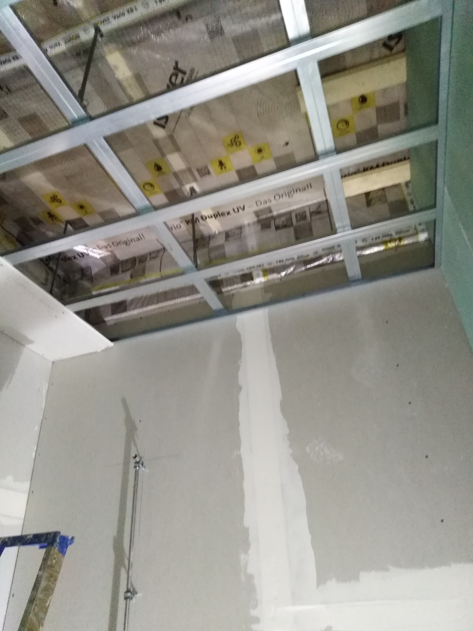 Metalowy stelaż sufitowy z izolacją, widoczne płyty gipsowe na ścianach w trakcie montażu, fragment drabiny malarskiej w lewym dolnym rogu.