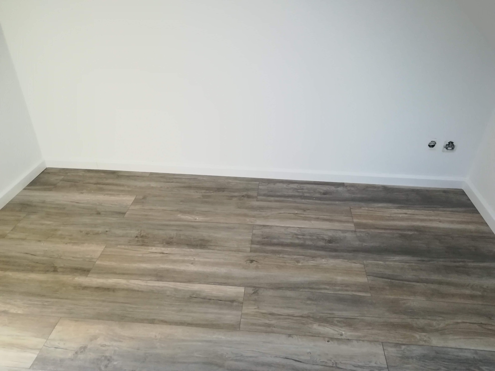 Ułożona podłoga z paneli imitujących drewno w jasnych szaro-beżowych odcieniach, widoczne gniazdka elektryczne na białej ścianie w rogu pomieszczenia.