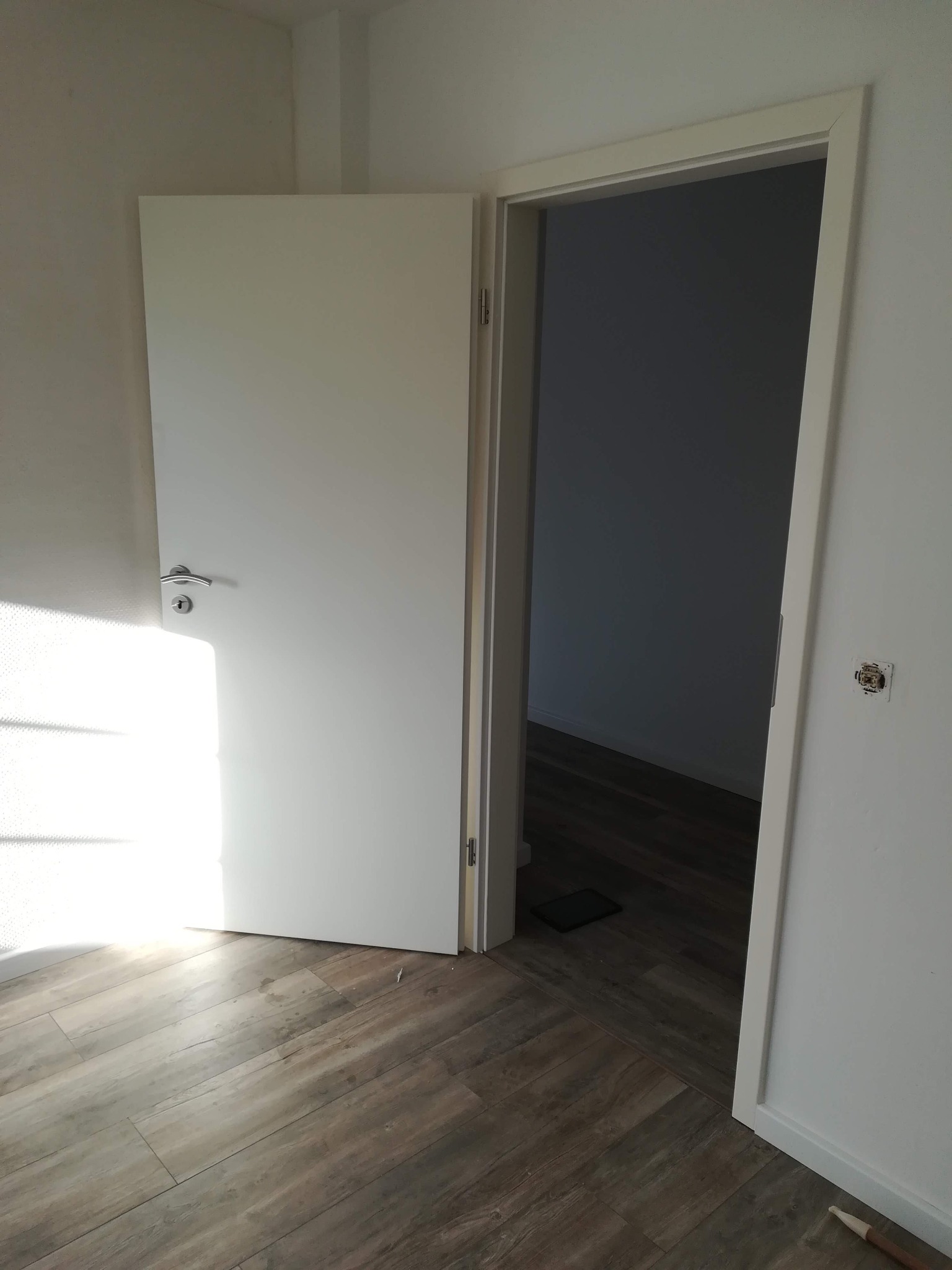 Białe drzwi wewnętrzne z chromowaną klamką, częściowo otwarte, z widocznym wnętrzem pomieszczenia z drewnopodobną podłogą; na ścianie widoczne gniazdko elektryczne w trakcie montażu.