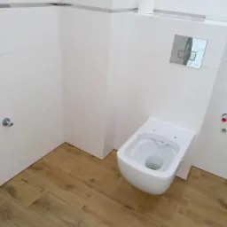 Nowoczesna toaleta podwieszana z chromowanym przyciskiem spłukującym, białe płytki ścienne z dekoracyjnym paskiem i podłoga imitująca drewno.