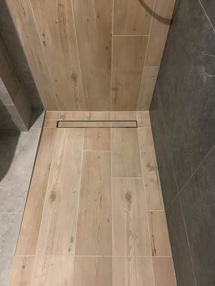 Wykończona kabina prysznicowa z odpływem liniowym, wyłożona płytkami imitującymi drewno na podłodze i ścianie oraz szarymi płytkami na jednej ze ścian.