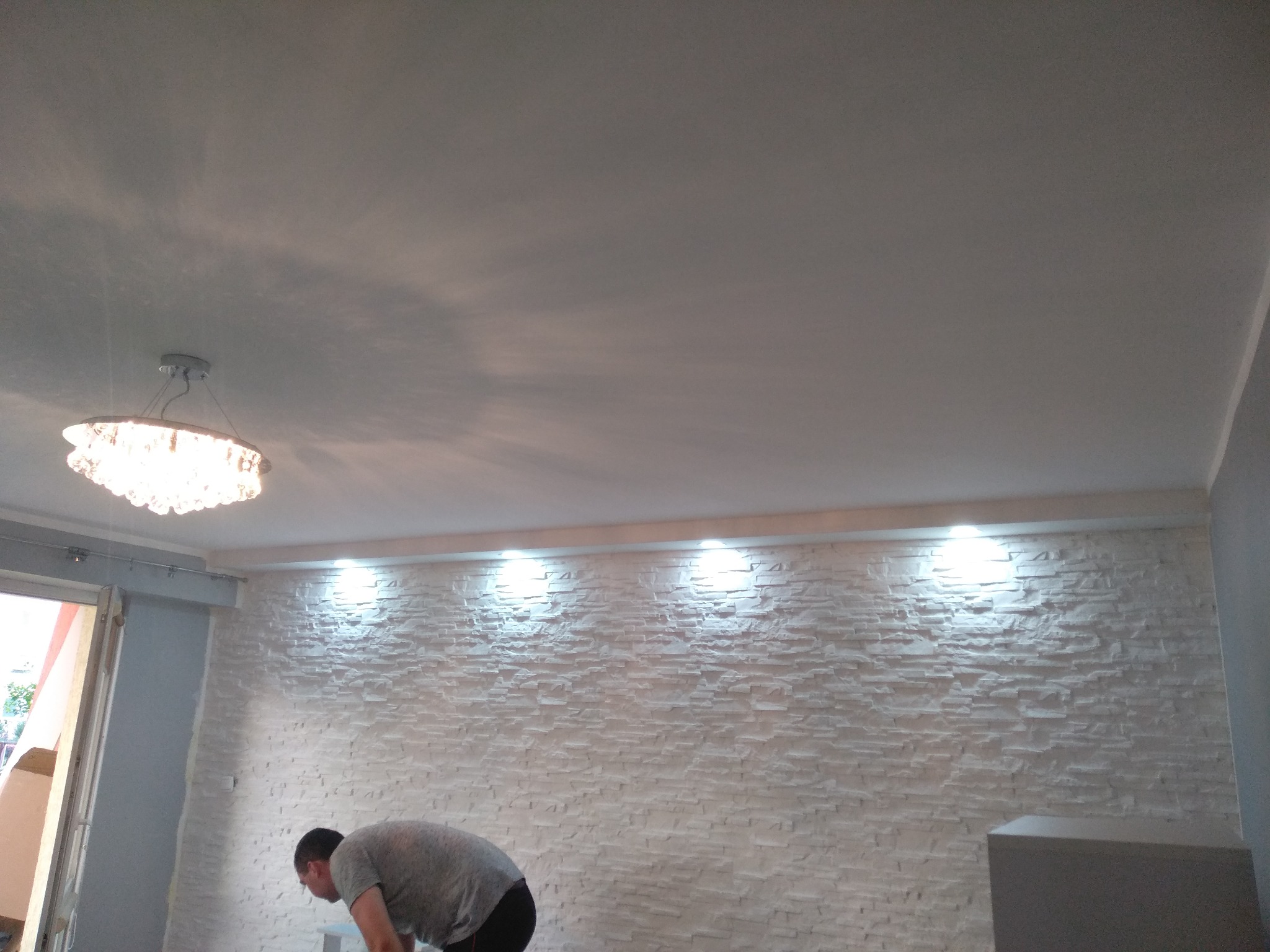 Montaż dekoracyjnej ściany z białego kamienia, oświetlonej czterema punktowymi lampami, z widocznym pracownikiem w trakcie pracy i kryształowym żyrandolem w tle.