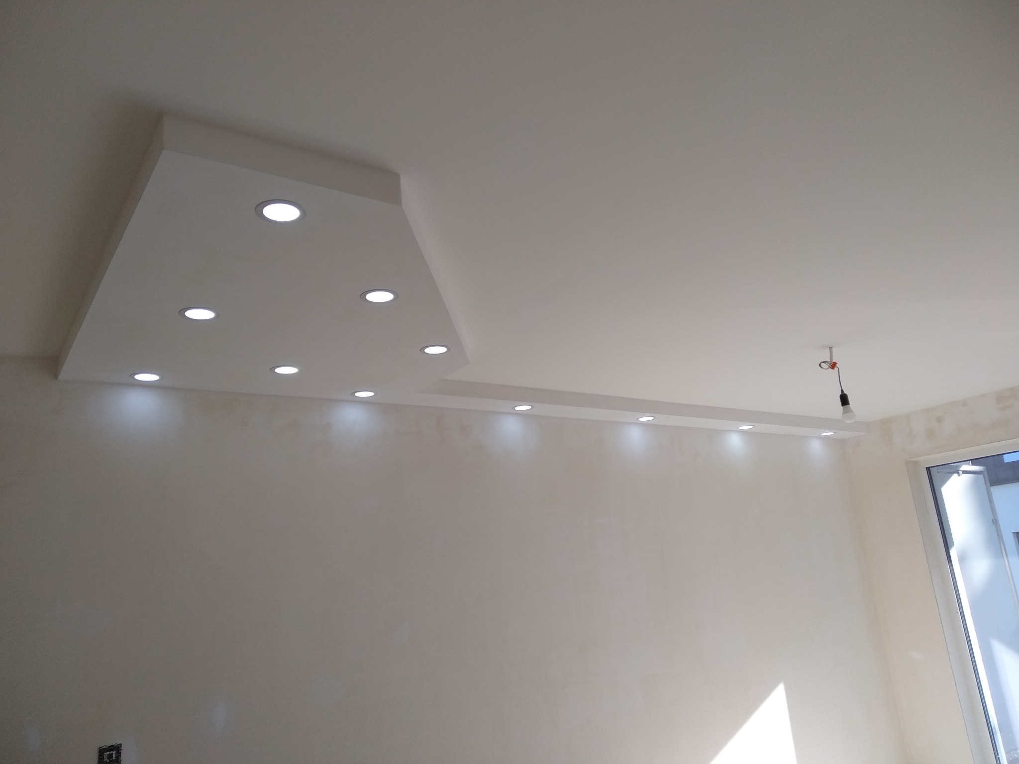 Nowoczesny sufit podwieszany z oświetleniem punktowym LED w jasnym pomieszczeniu, widoczny fragment okna i niedokończone wykończenie ściany.