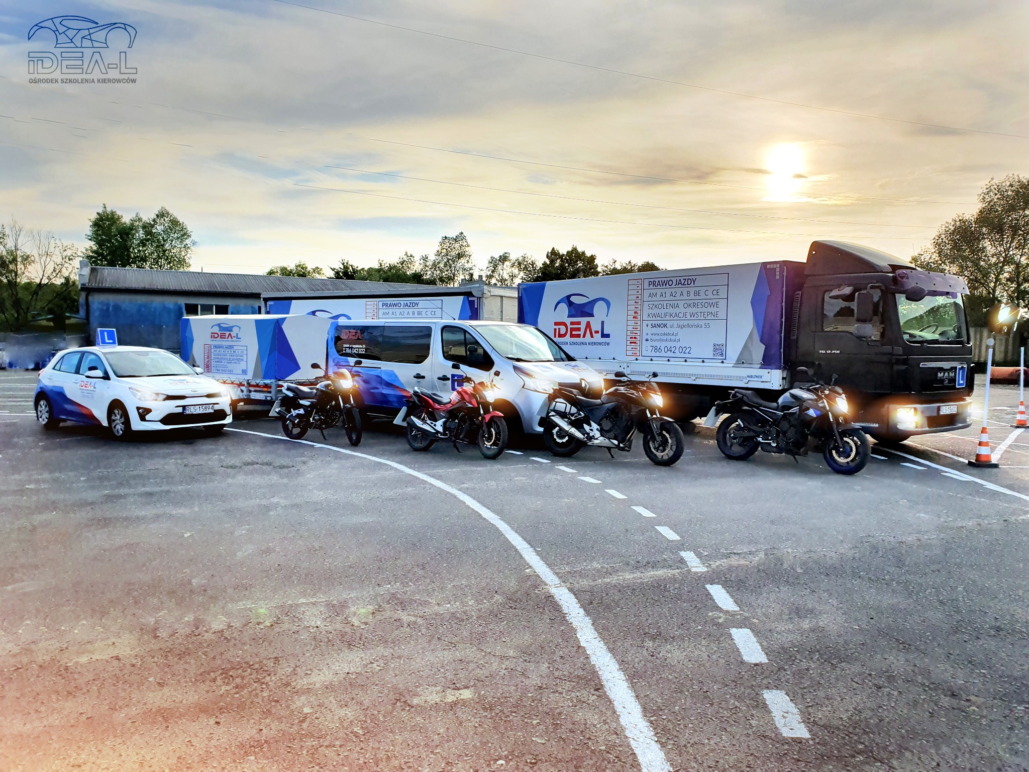 Flota pojazdów: samochód osobowy, furgonetka, ciężarówka i motocykle, oznakowane logo ośrodka szkolenia kierowców, ustawione na placu manewrowym z namalowanymi liniami, w tle budynek z napisem 'L'.