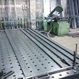 F-tech - Stół spawalniczy z perforowanym blatem, na którym zamocowane są metalowe profile za pomocą ścisków, w tle piła taśmowa do metalu i stosy metalowych elementów.