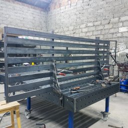 F-tech - Konstrukcja metalowa z profili, prawdopodobnie rama lub podstawa, spawana na stole spawalniczym w warsztacie, widoczne narzędzia i butla z gazem.