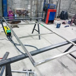 F-tech - Warsztatowa sceneria z ramą ze stali nierdzewnej na kozłach, obok spawarka, butle z gazami technicznymi i narzędzia ślusarskie, surowe wnętrze z betonowymi ścianami.