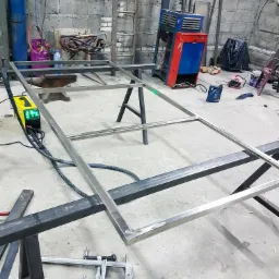 Warsztatowa sceneria z ramą ze stali nierdzewnej na kozłach, obok spawarka, butle z gazami technicznymi i narzędzia ślusarskie, surowe wnętrze z betonowymi ścianami.