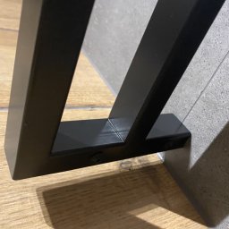 F-tech - Fragment czarnej, metalowej konstrukcji, prawdopodobnie nogi stołu lub innego mebla, przymocowanej śrubami do podłogi i ściany wyłożonej płytkami.