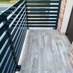 F-tech - Nowoczesna, pozioma, czarna konstrukcja na balkonie z płytkami imitującymi drewno, mocowana do ceglanej ściany budynku.