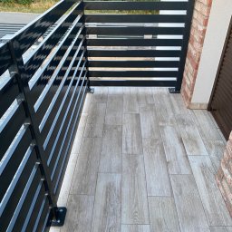 F-tech - Nowoczesny balkon z czarną, metalową balustradą o poziomych szczeblach, wykończony płytkami imitującymi drewno i fragmentem ściany z cegły.