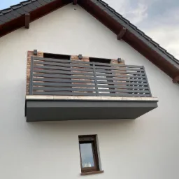 Szara, pozioma balustrada balkonowa z prostokątnych profili na tle ceglanej wstawki w białej elewacji domu jednorodzinnego, widok z dołu, małe okno poniżej.