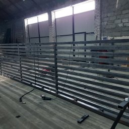 F-tech - Surowa, metalowa konstrukcja z poziomych profili w warsztacie, widoczny spawacz i szlifierka kątowa, ceglana ściana w tle.