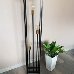 F-tech - Czarna, metalowa lampa podłogowa w stylu industrialnym z trzema żarówkami Edisona, stojąca obok białej doniczki z zieloną rośliną, na szarych panelach podłogowych.