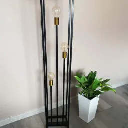 Czarna, metalowa lampa podłogowa w stylu industrialnym z trzema żarówkami Edisona, stojąca obok białej doniczki z zieloną rośliną, na szarych panelach podłogowych.