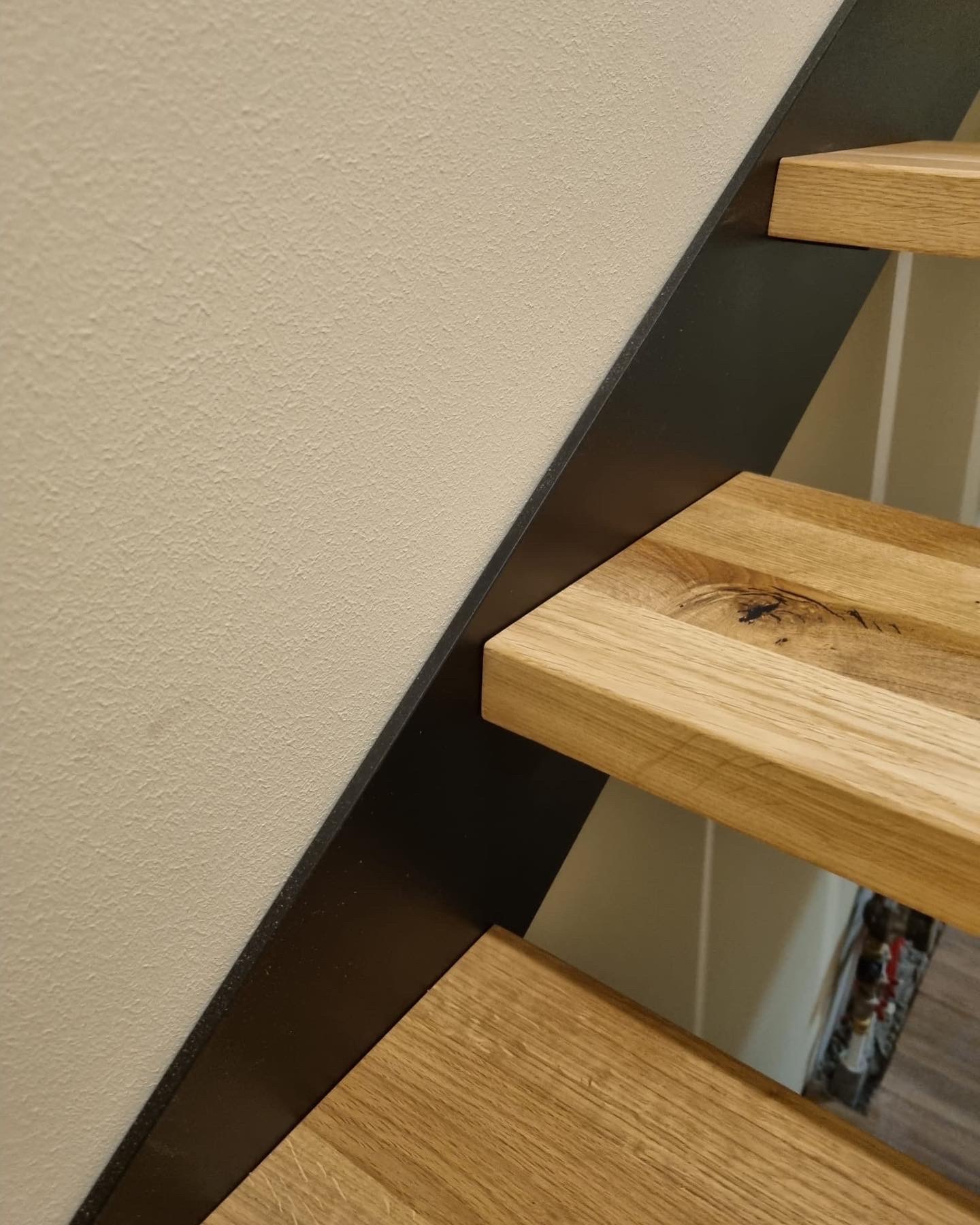 Fragment schodów z drewnianymi stopniami i czarną metalową konstrukcją nośną, widoczny fragment ściany w tle.