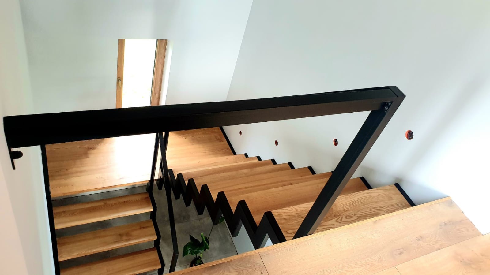 Nowoczesne schody drewniane z czarną, geometryczną konstrukcją nośną i poręczą, widok z góry w jasnym wnętrzu.