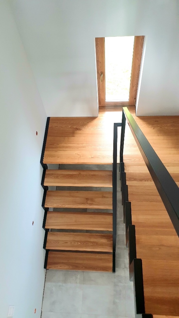 Nowoczesne schody drewniane z czarną metalową konstrukcją, widok z góry, jasne wnętrze z oknem w tle.