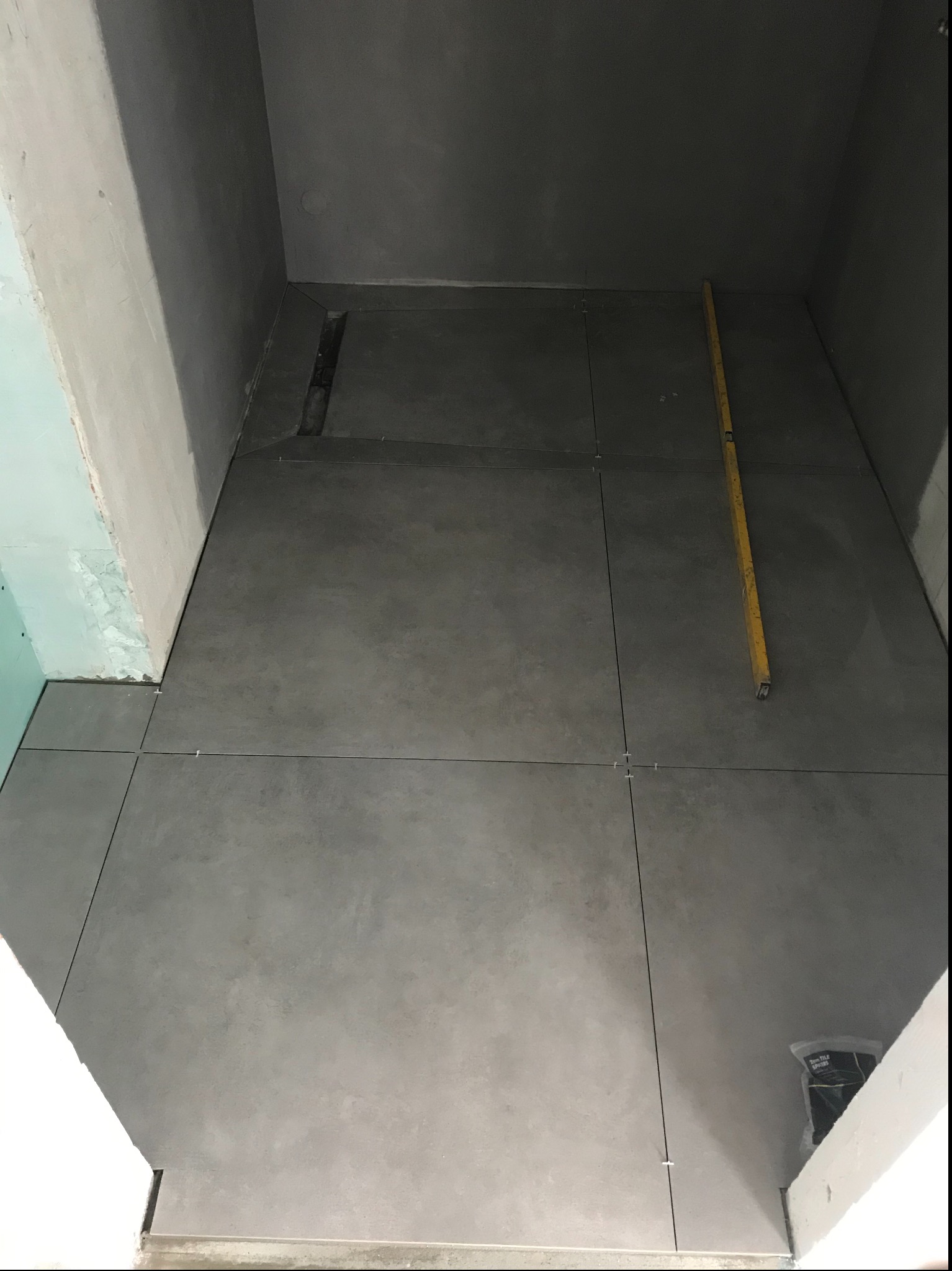 Szara, matowa podłoga z płytek w trakcie układania, widoczny odpływ liniowy, poziomica i opakowanie fugi.