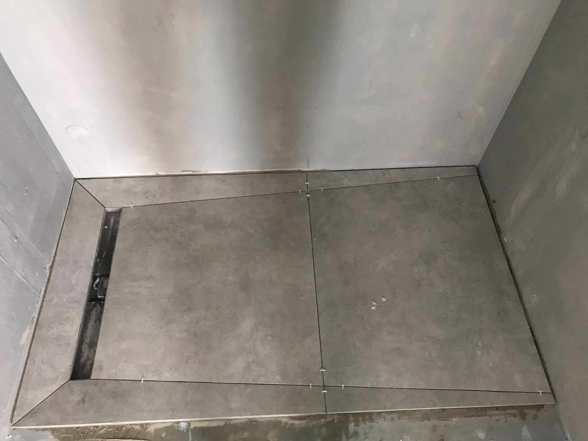 Szare płytki podłogowe w trakcie układania w łazience, widoczny odpływ liniowy i krzyżyki dystansowe.