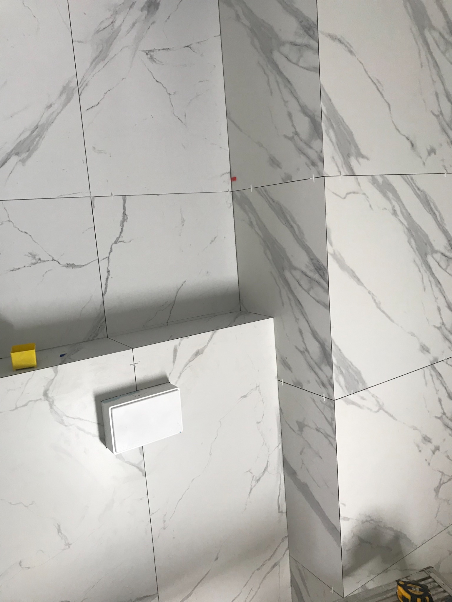 Montaż płytek imitujących marmur na ścianie z użyciem krzyżyków dystansowych; widoczny fragment ściany, podłogi i obudowy, żółty pojemnik i miara zwijana w rogu kadru.
