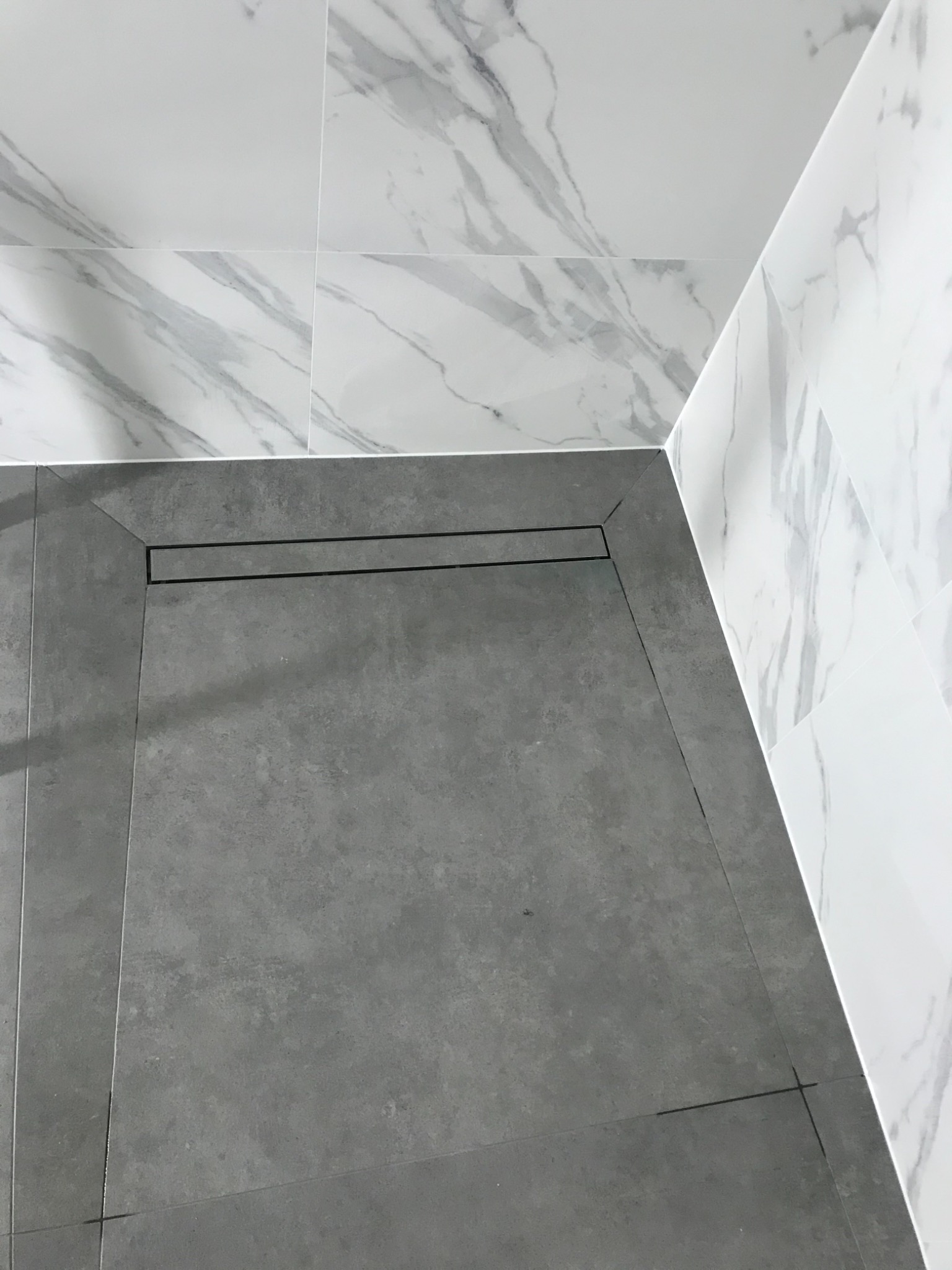 Szara, matowa podłoga wyłożona płytkami w kabinie prysznicowej z liniowym odpływem; ściany wykończone białymi płytkami imitującymi marmur.