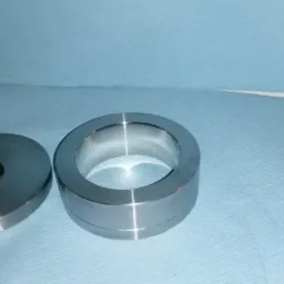 Dwie precyzyjnie obrobione, metalowe części: okrągła płytka z otworem i pierścień, leżące na niebieskim tle.