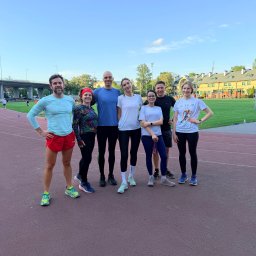 GetThatSpeed - Gabriel Mikołajewski - Grupa siedmiu osób w strojach sportowych pozuje na bieżni stadionu, z budynkiem w tle i błękitnym niebem. Uśmiechnięci, gotowi do treningu biegowego.