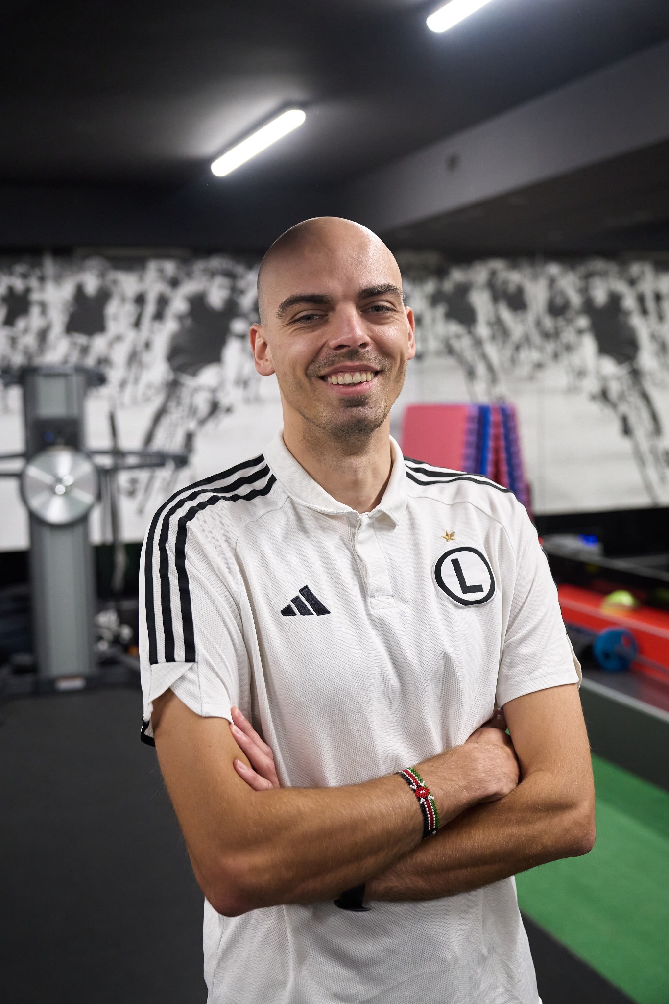 Uśmiechnięty mężczyzna w koszulce sportowej z logo klubu, stojący na tle sali treningowej z maszynami i grafiką na ścianie, z bransoletką na ręce.