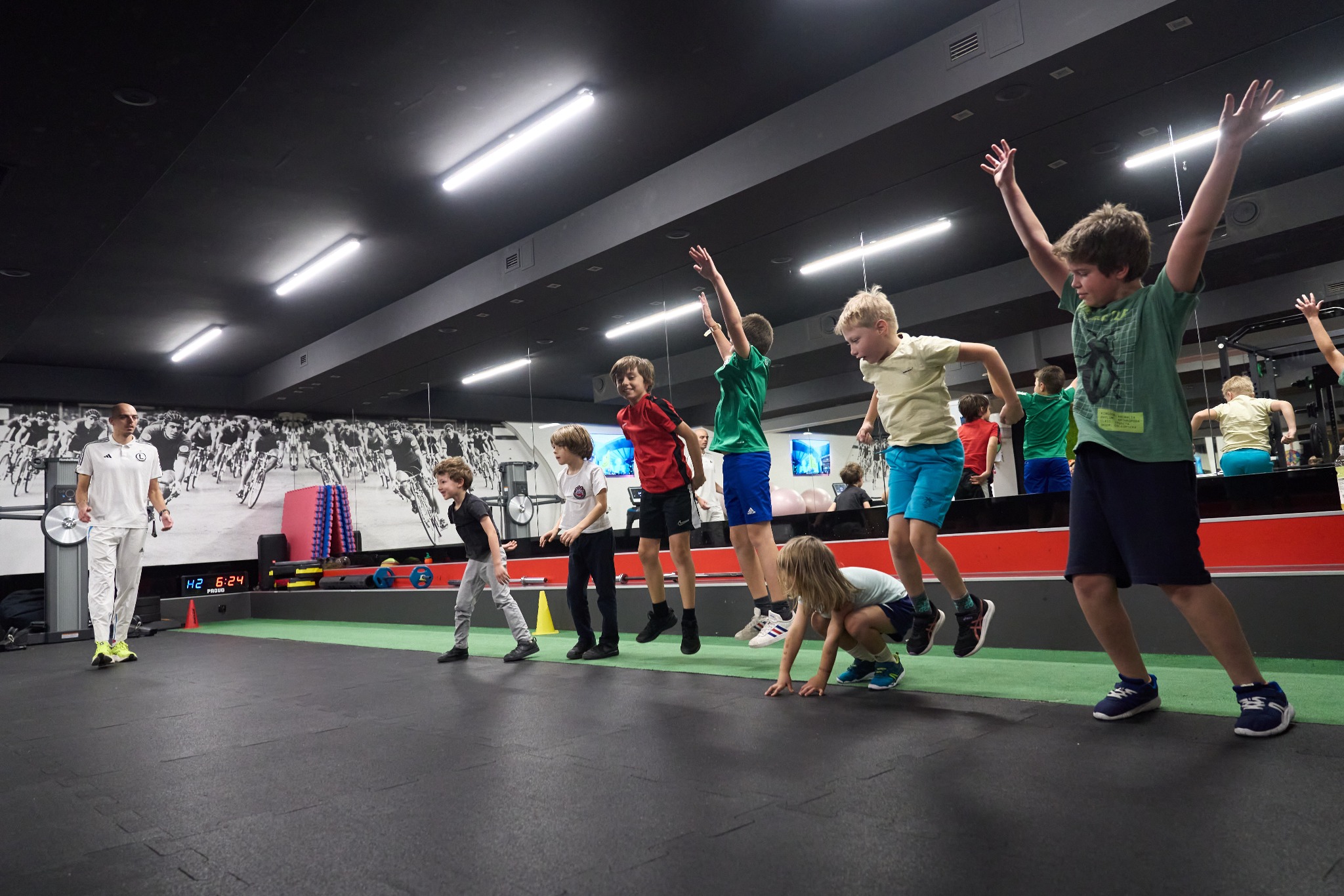 Grupa dzieci wykonuje ćwiczenia ogólnorozwojowe pod nadzorem trenera w sali gimnastycznej z motywem kolarskim na ścianie.