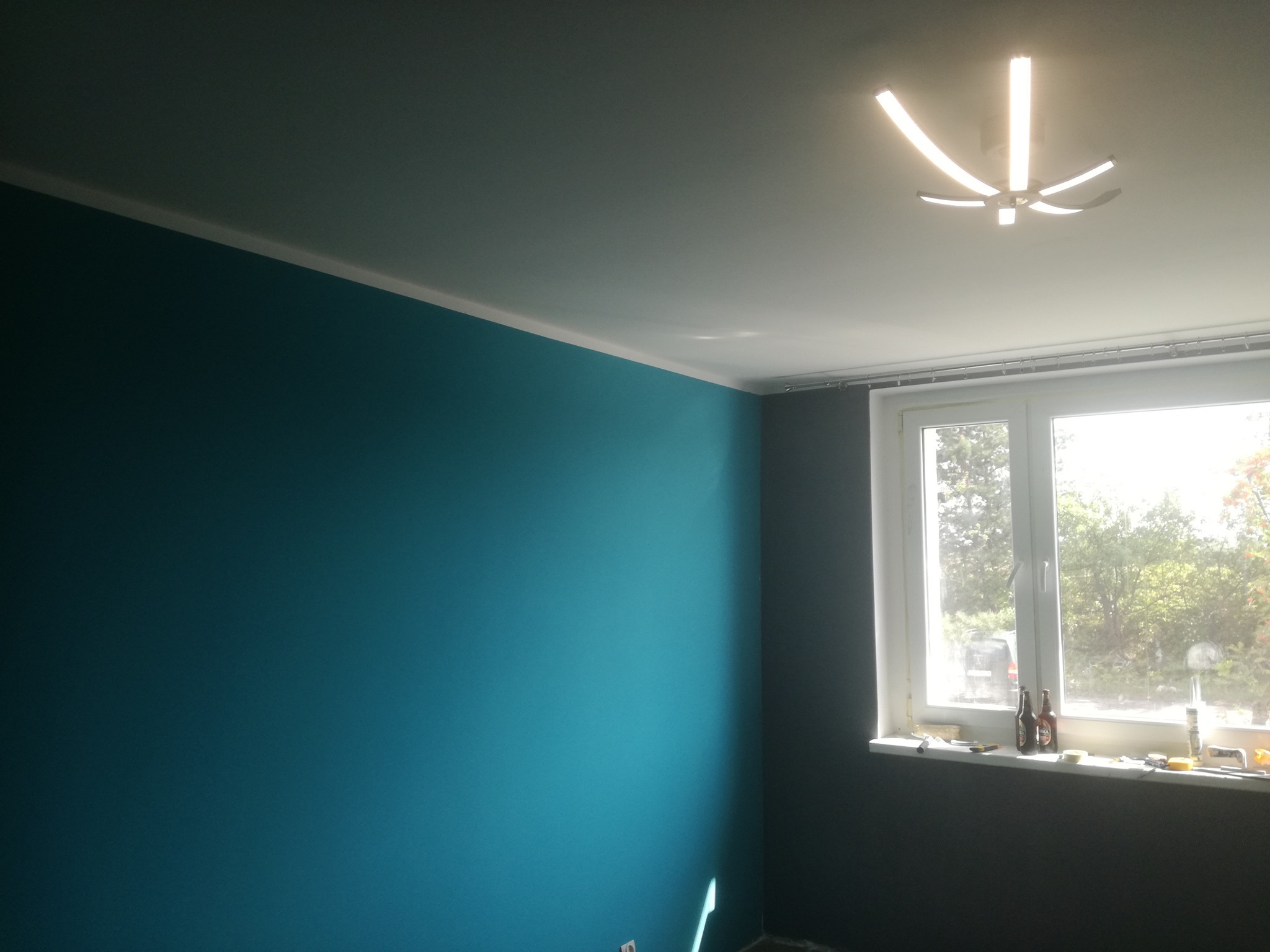 Pomieszczenie po malowaniu: ściana w kolorze turkusowym z białym sufitem i listwą, widoczne okno z narzędziami i butelkami na parapecie, nowoczesna lampa sufitowa.