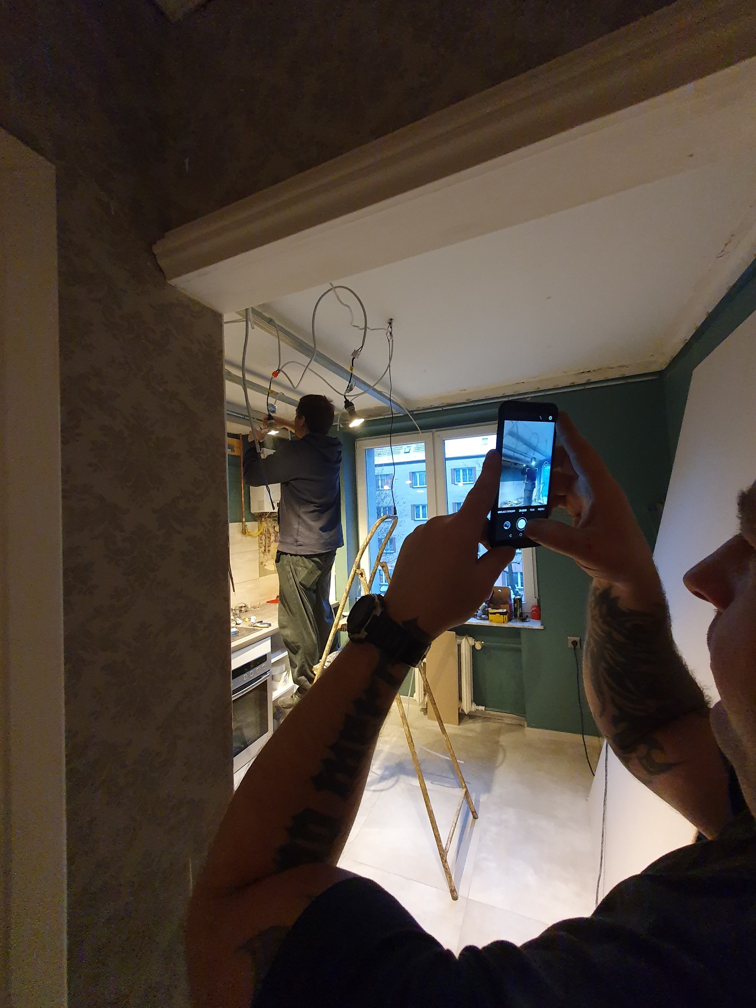 Elektryk na drabinie montuje oświetlenie pod sufitem w trakcie remontu, fotografowane telefonem przez drugą osobę z bliska, widoczne kable, okno i fragmenty pomieszczenia.