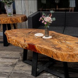 Studio Obiektyw - Dwa stoliki kawowe z plastrów drewna na czarnych, metalowych nogach, jeden z bukietem kwiatów, na drewnianym tarasie obok czarnej sofy.