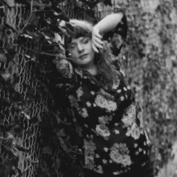 Portret kobiety w kwiecistej bluzce, opierającej się o zarośnięte ogrodzenie z siatki w czarno-białej tonacji.