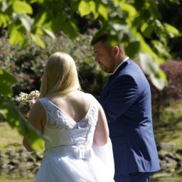 Studio Obiektyw - Para młoda w plenerze: panna młoda w białej sukni z bukietem, pan młody w granatowym garniturze, zielone tło z drzewami i krzewami, ujęcie zza liści.