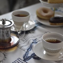 Studio Obiektyw - Szklany czajnik z zaparzaczem, filiżanki z herbatą i talerz z pączkami na wzorzystym obrusie w neutralnej tonacji.